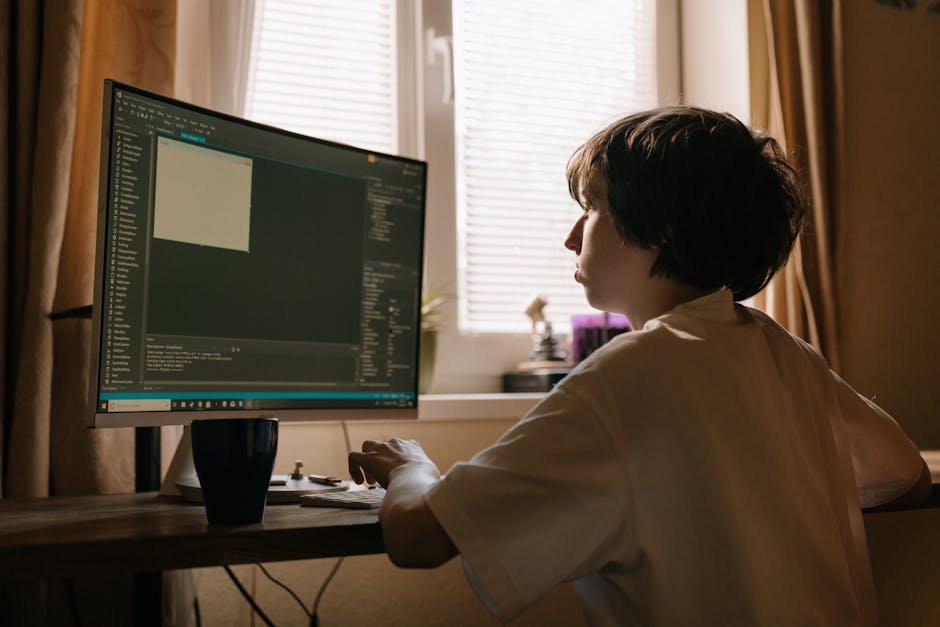 A woman with short, dark hair is seated at a wooden desk indoors, working on a desktop computer with a large monitor displaying a dark-themed code editor or design software. The room is illuminated by