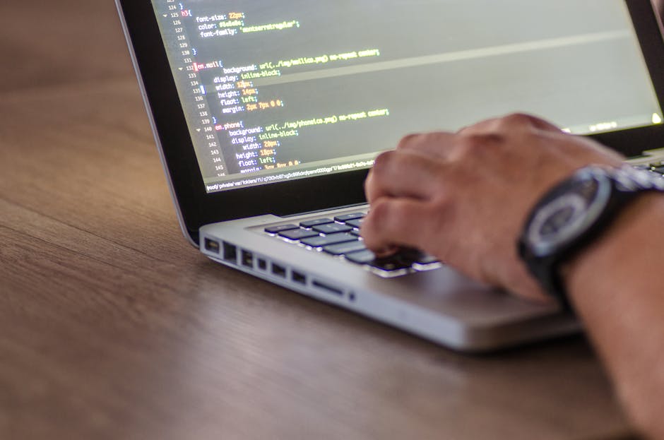 A close-up image of a person's hand operating a laptop computer on a wooden surface, with visible code displayed on the screen. The individual is wearing a black wristwatch, and the scene is lit with 