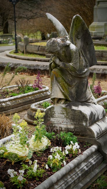 A peaceful section of a cemetery garden featuring weathered stone headstones of various shapes and sizes standing amidst dense green foliage. The headstones display signs of moss and age, with some le